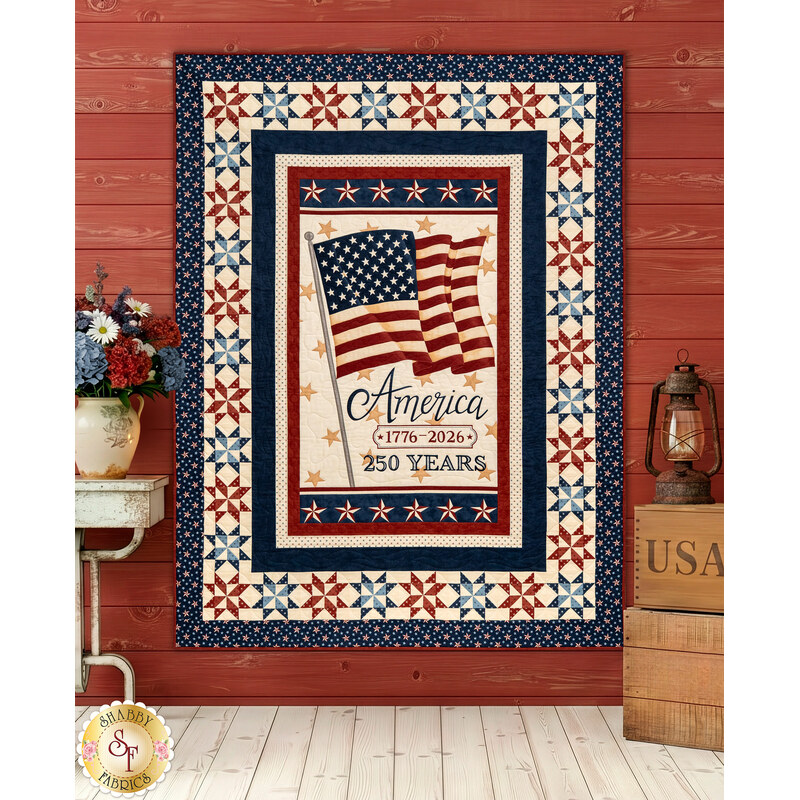 The completed America 250 Patchwork Panel Quilt colored in red, navy, and cream fabrics from the America 250 collection by Moda Fabrics, staged on an antiqued red wall beside coordinating rustic décor.