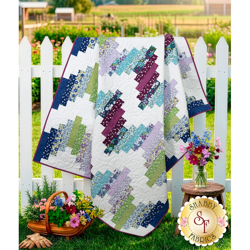 The completed Ridiculously Easy Jelly Roll Quilt in fabric from the Seaside Garden collection, staged on a white picket fence with wooden stool, cut flowers, and gardening gloves.