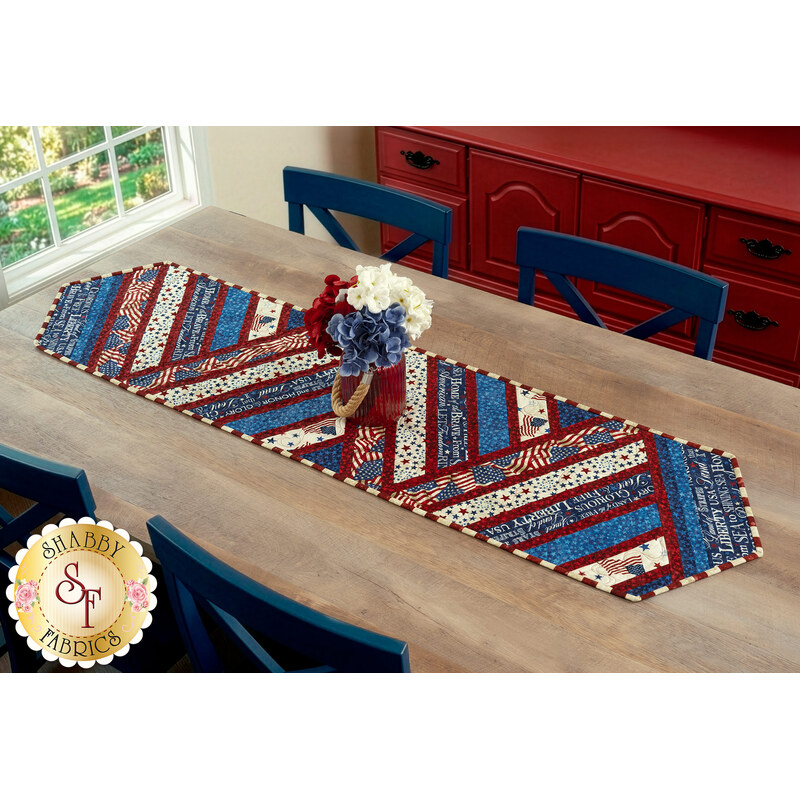 The completed Greased Lightning Table Runner in red, white, and blue fabrics from the Stars & Stripes 14 collection by Northcott Fabrics, staged on a rustic wood table with flowers.