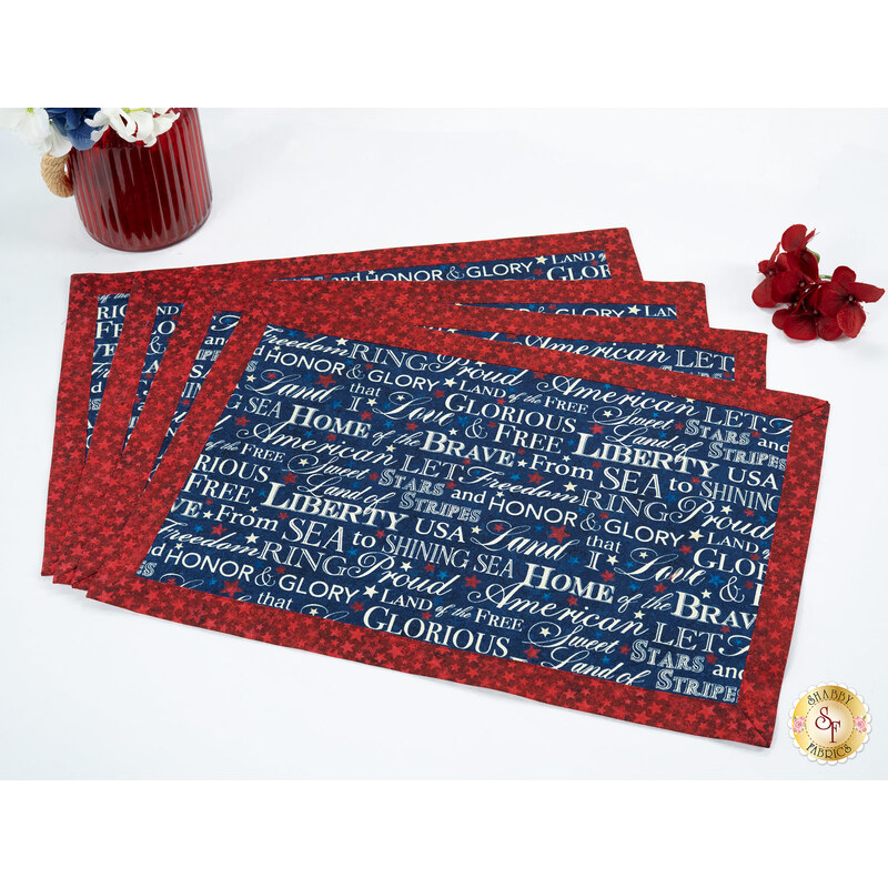 Four Self-Binding Placemats with a navy blue print featuring patriotic words and phrases, framed by a crimson border fabric, staged on a white table with coordinating décor.