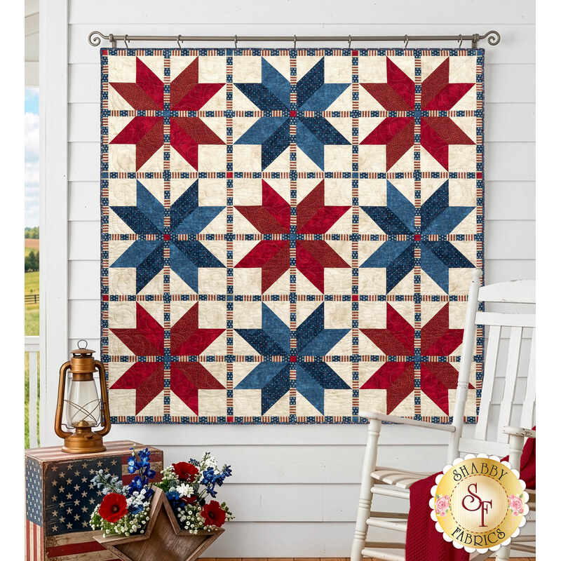 The completed Vintage Stars Quilt, colored in red, cream, and blue fabrics from the American Heartland collection Northcott Fabrics, hung on a white paneled wall on a porch and staged with coordinating décor.
