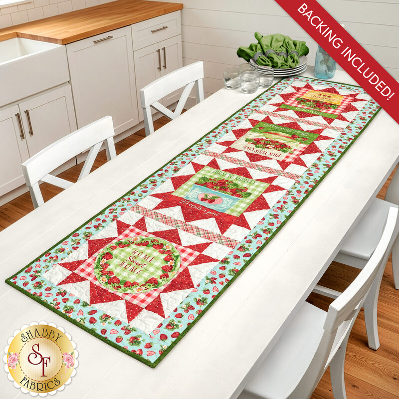 The completed Double Geese Table Runner, colored in aqua, green, white, and red fabrics from the Berry Besties collection by Henry Glass Fabrics, staged on a white kitchen table with coordinating table settings at one end.