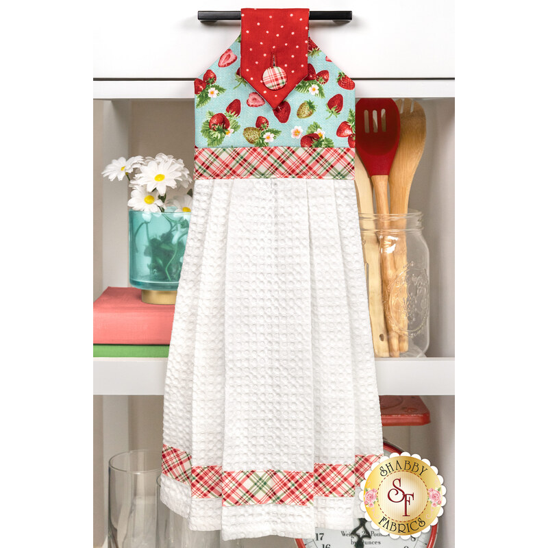 The completed Hanging Towel colored in aqua and red fabrics from the Berry Besties collection by Henry Glass Fabrics, hung on a white kitchen drawer and staged with coordinating décor.