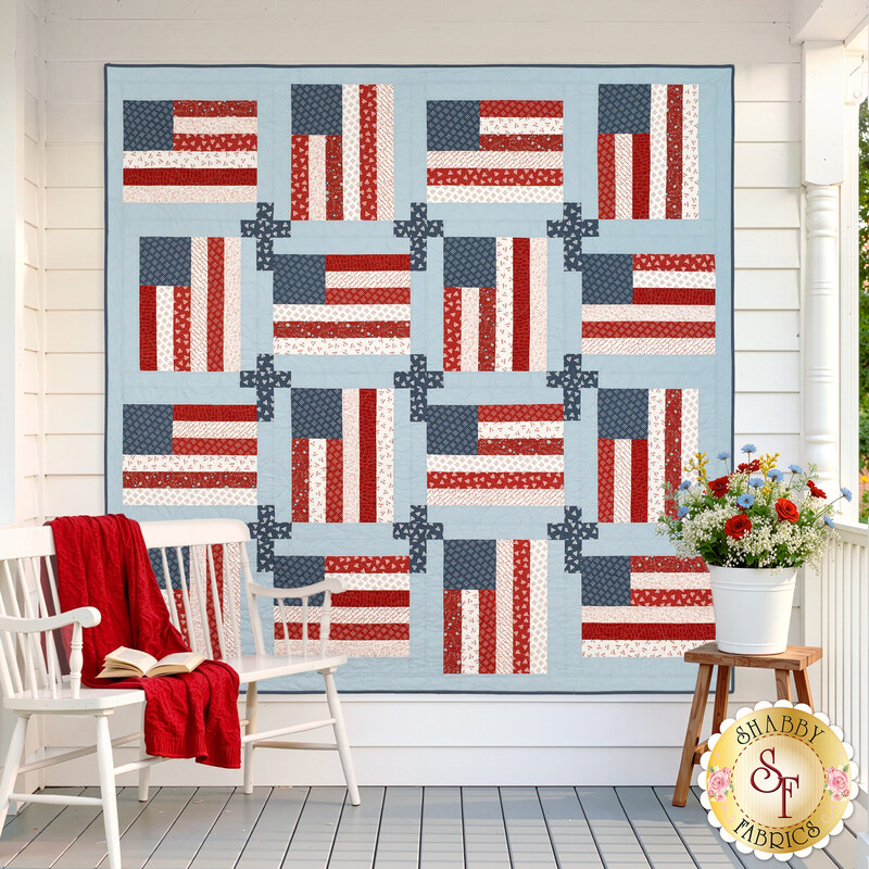 The completed God and Country Quilt featuring alternating red, white, and blue fabrics from the Independence Day collection from Moda Fabrics hangs on a porch, with a chair and flowers nearby.