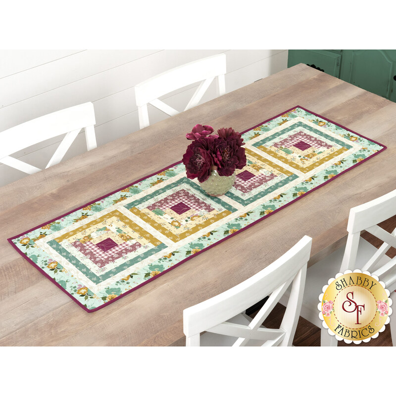 The completed table runner made with log cabin blocks of plum purple, ocher, and teal floral fabrics from the Renew collection, laying in the center of a wooden dining table with white chairs.