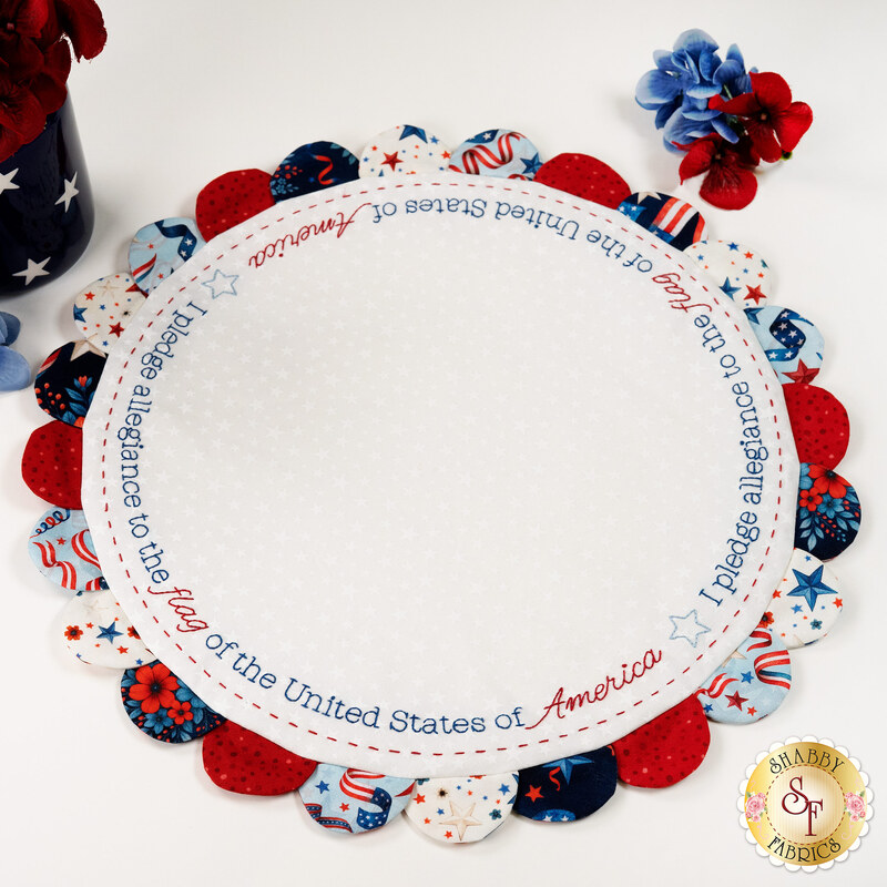 Circular fabric table topper with scallops in patriotic colors and the embroidered text, "I pledge allegiance to the flag," staged on a white table with coordinating décor.