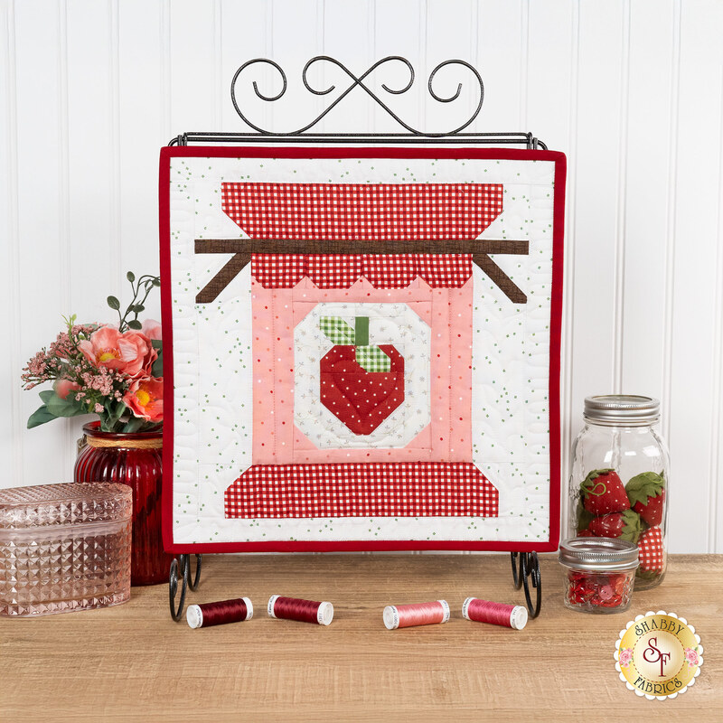 The completed A Year in Spools - June - Sew Jammy Kit, colored in white, red, and pink fabrics, staged on a display stand with coordinating décor.