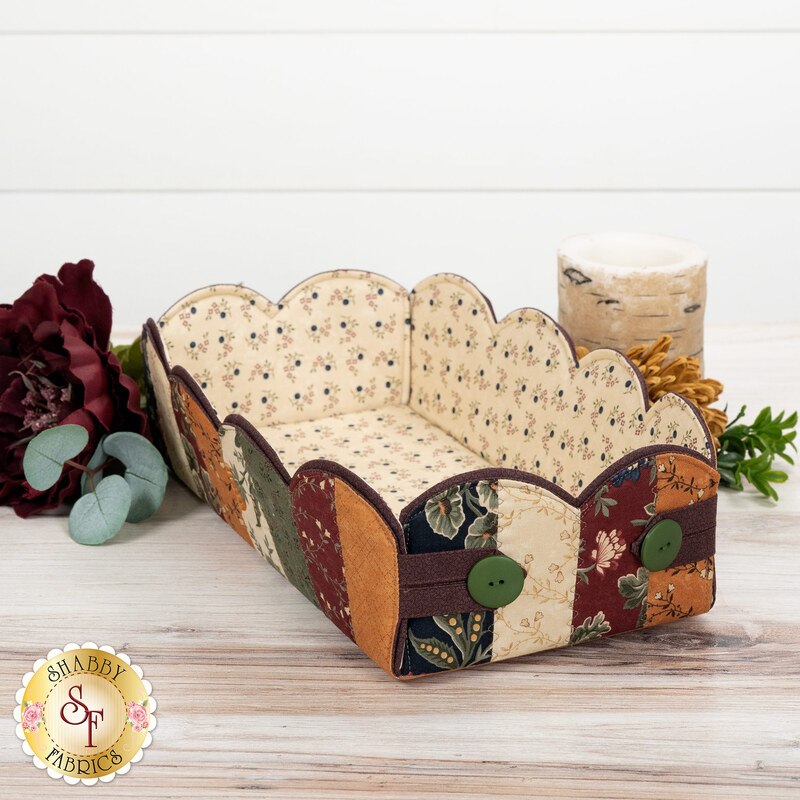 The completed Scalloped Basket, colored in primitive fabrics from the Quail Run collection by Moda, staged on a wooden table with coordinating flowers and décor.