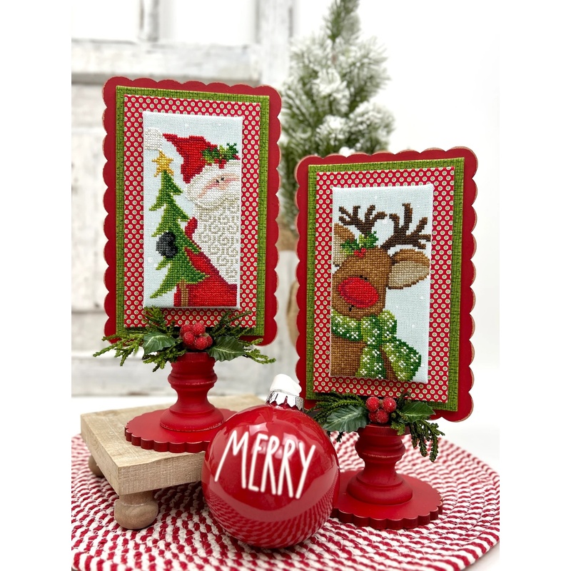 The two completed cross stitch projects of Santa & Rudolph, set on pale blue fabric and stitched with bright colors, displayed on a Paisleys and Polka Dots mount painted red and green.