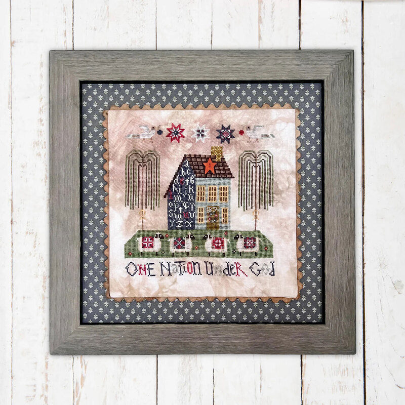 The framed cross stitch project featuring a house, stars, and the phrase One Nation Under God.