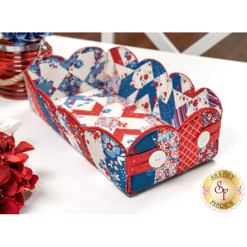 The completed Scalloped Basket, colored in red, white, and blue fabrics, staged on a white table with coordinating flowers and décor.