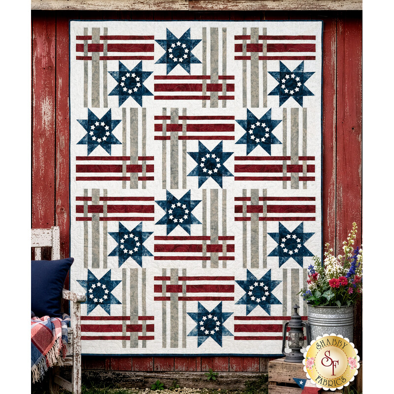 The completed Hometown Honor quilt, a geometric stripe and star design colored in gray, white, red, and blue batik fabrics from the Missing You collection from Island Batik, staged on a red barn wall with coordinating décor.