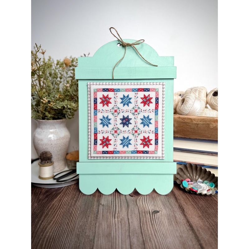 A mint-framed cross-stitch artwork sits on a wooden table, surrounded by a vase and buttons.