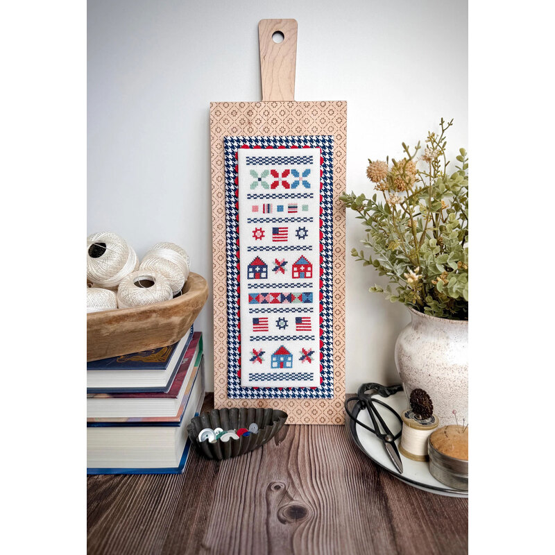 The finished Hometown Summer cross stitch project mounted on a wooden cutting board, surrounded by books, thread spools, and a plant.