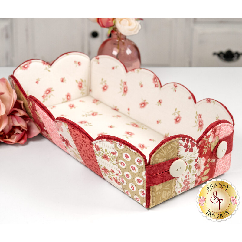 The completed Scalloped Basket, colored in green, white, pink, and red fabrics from the Elizabeth collection by Studio E, staged on a white table with coordinating flowers and décor.