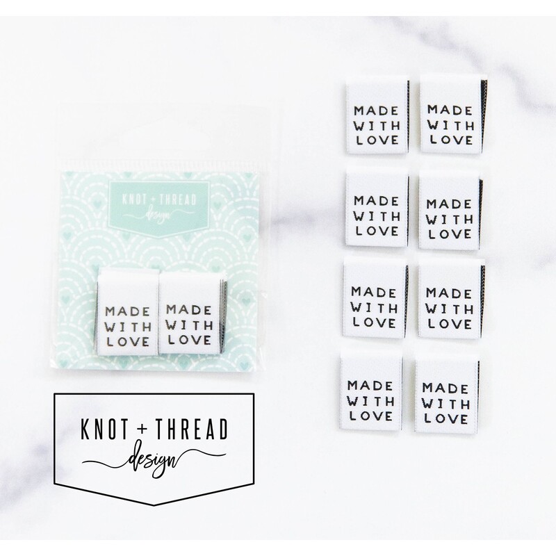 8 "Made with Love" fabric tags lined up and in packaging by Knot and Thread Designs on a white marble background. 