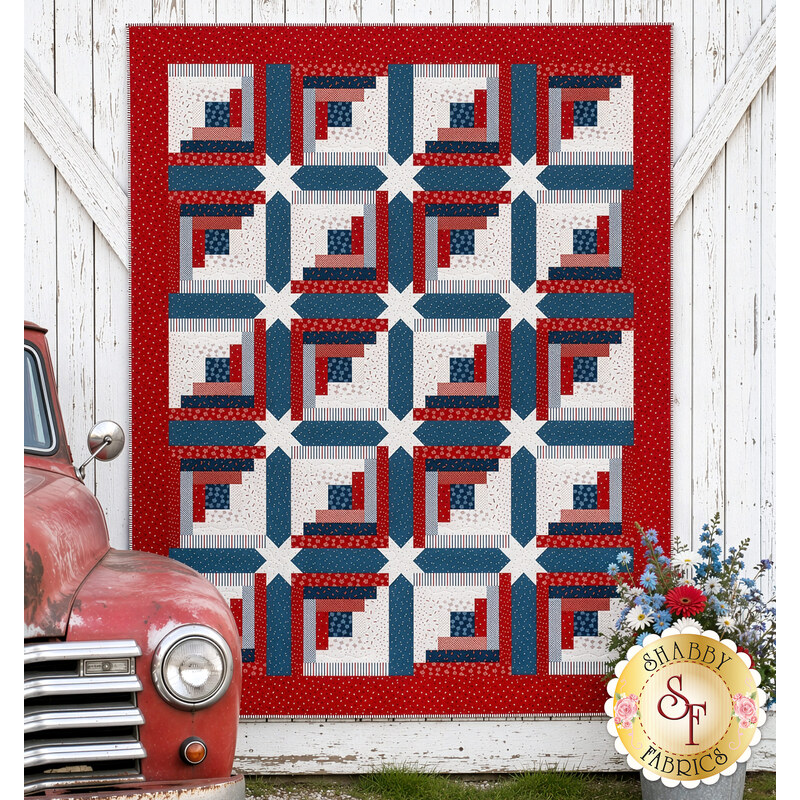 The completed Steadfast Quilt, colored in red, white, and blue fabrics from the Seasonal Basics collection by Riley Blake Designs, staged on a white barn door wall and staged with flowers and a vintage red truck.
