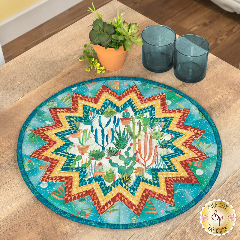 A round table topper with a bursting star design in vibrant southwest inspired fabrics from the Desert Dreams collection by Maywood Studio, staged on a wooden table with coordinating décor.