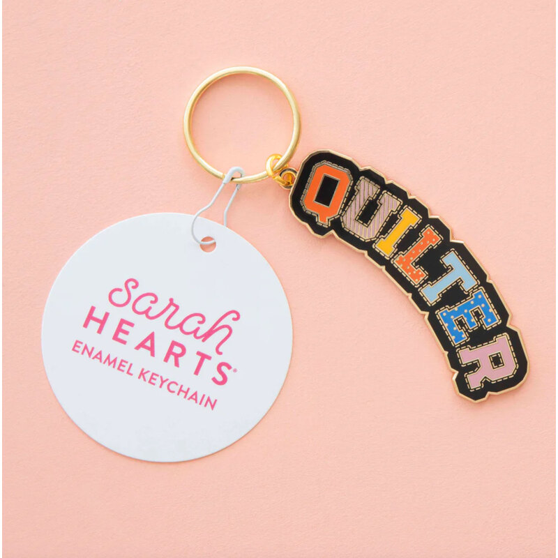 Enamel keychain featuring the word quilter spelled out with stitching, isolated on a pink background.
