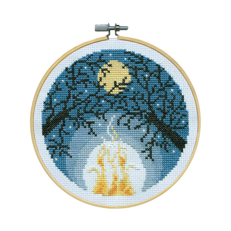 A cross-stitched night sky scene with shadowed tree branches and a fire below, framed in a wooden embroidery hoop.