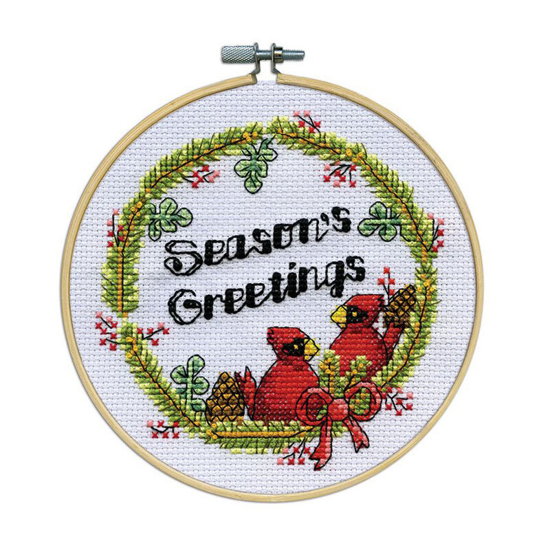 A cross-stitched "Season's Greetings" with 2 red cardinals framed by a berry and pine leaf wreath, framed in a wooden embroidery hoop.