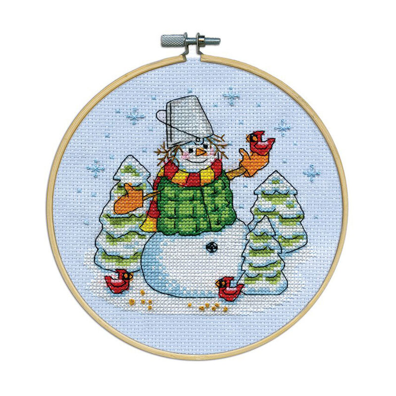 A cross-stitched bundled up snowman wearing a bucket hat, surrounded by trees, birds, and blue snowflakes, framed in a wooden embroidery hoop.