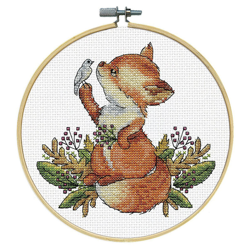 The completed cross stitch fox holding a bird nestled in leaves and berries, framed in a wooden embroidery hoop.