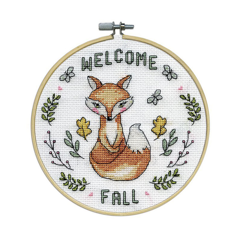 The completed cross stitch featuring the words Welcome Fall and a fox surrounded by leaves and butterflies, framed in a wooden embroidery hoop.