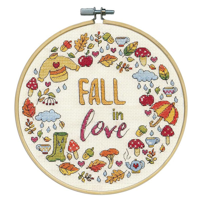 The completed cross stitch featuring the words "Fall in love" surrounded by fall themed motifs , framed in a wooden embroidery hoop.