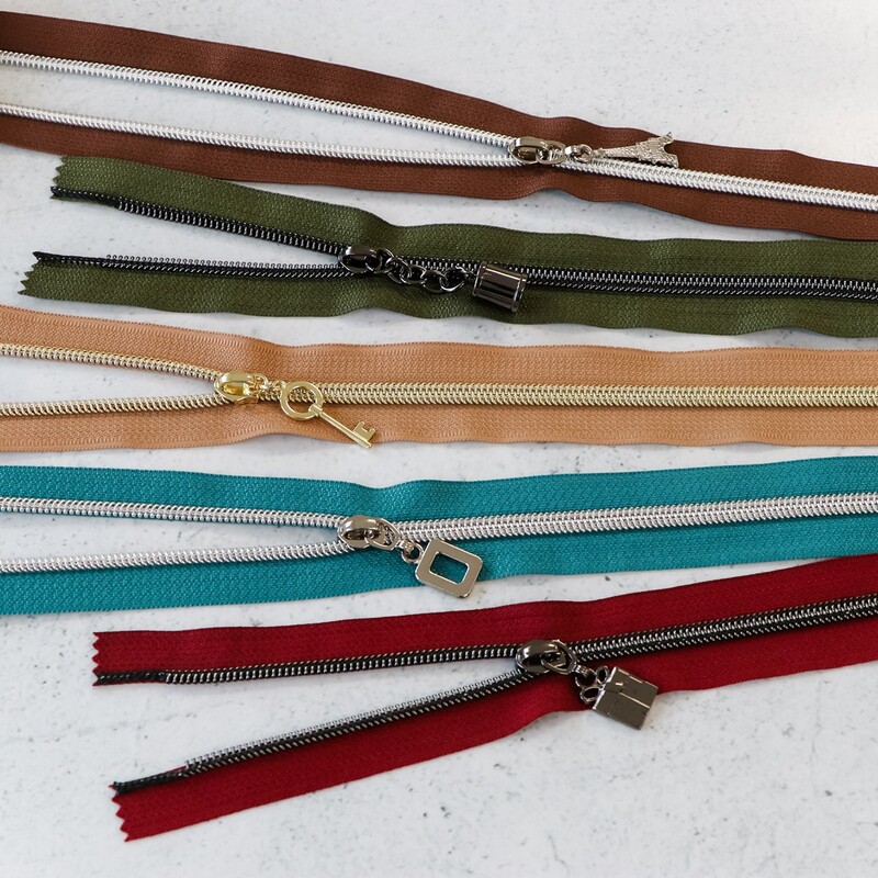 Five zipper pieces in various colors arranged horizontally, showing their coordinating novelty zipper pulls.