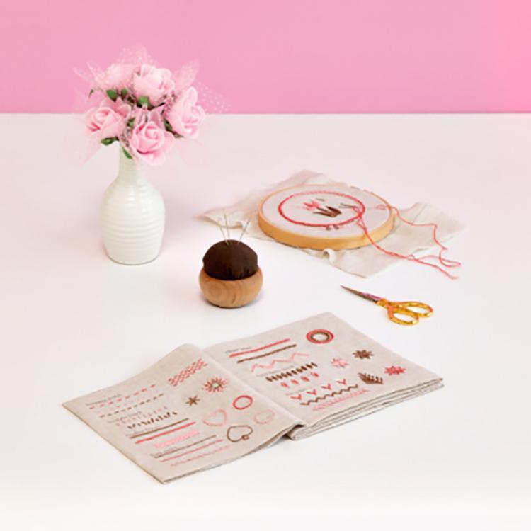 Open booklet of various pink stitches on a pink background with scissors, pin cushion, flowers, and a hoop.