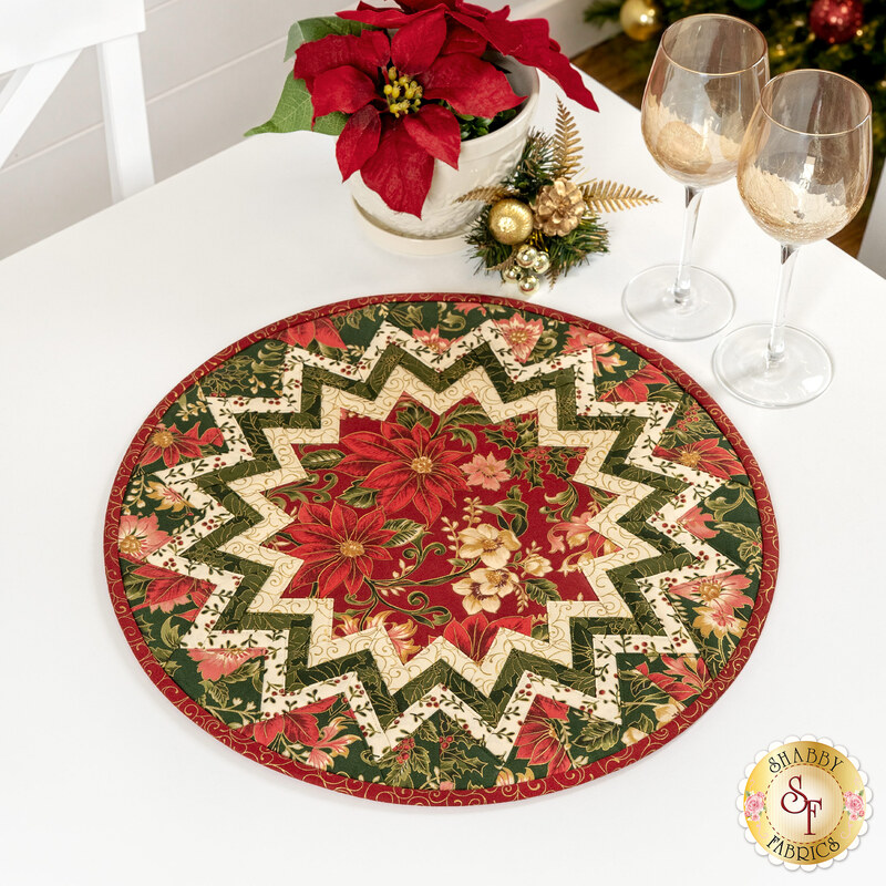 The completed topper, colored in red, cream, and green metallic fabrics from the Merry Manor Metallic 2025 collection from Moda Fabrics, staged on a white table with coordinating glassware and décor.