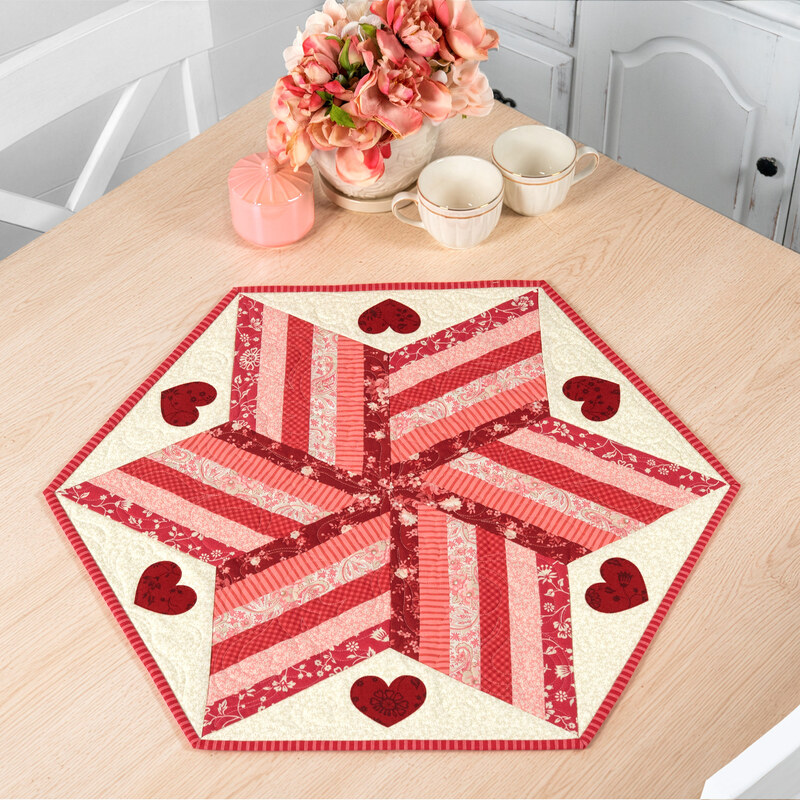 The completed 60 Degree Diamond Table Topper, colored in pink, red, and cream fabrics from the Petals and Polka Dots collection by Moda Fabrics, staged on a white table with coordinating flowers and teacups.