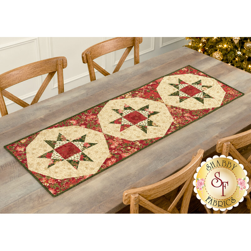The completed Any 12 Will Do Table Runner, colored in cream, green, and red metallic fabrics from the Merry Manor Metallic 2025 collection by Moda Fabrics, staged on a light wooden table with four chairs and a decorated Christmas tree peeking into frame from the upper right corner.