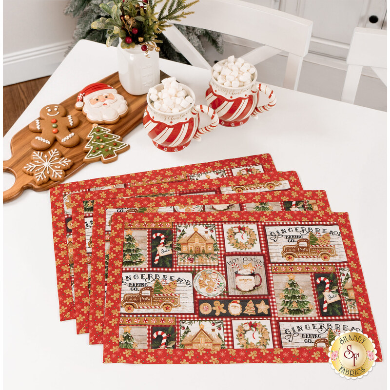 The four completed Self-Binding Placemats, fanned out and colored in red, black, beige, and green fabrics from the Very Merry Gingerbread collection by 3Wishes, staged on a white table with mugs of cocoa and adorable Christmas cookies.