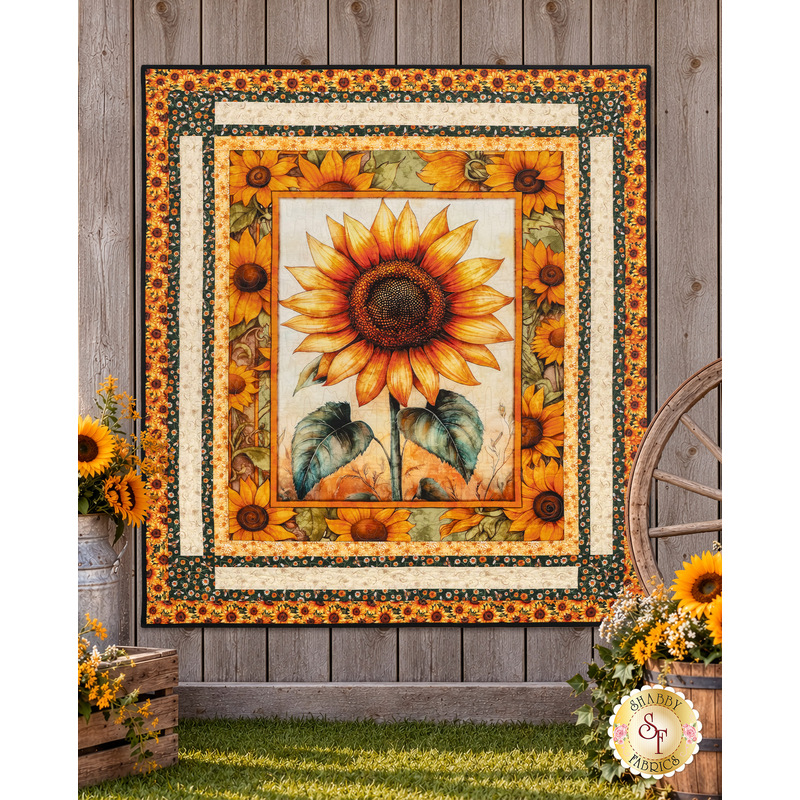 The completed Sunflower Serenade Panel Quilt, colored in cream, golden yellow, brown, and green fabrics from the Sunflower Serenade collection by QT Fabrics, hung on a rustic brown fence and staged with coordinating décor and baskets of sunflowers.
