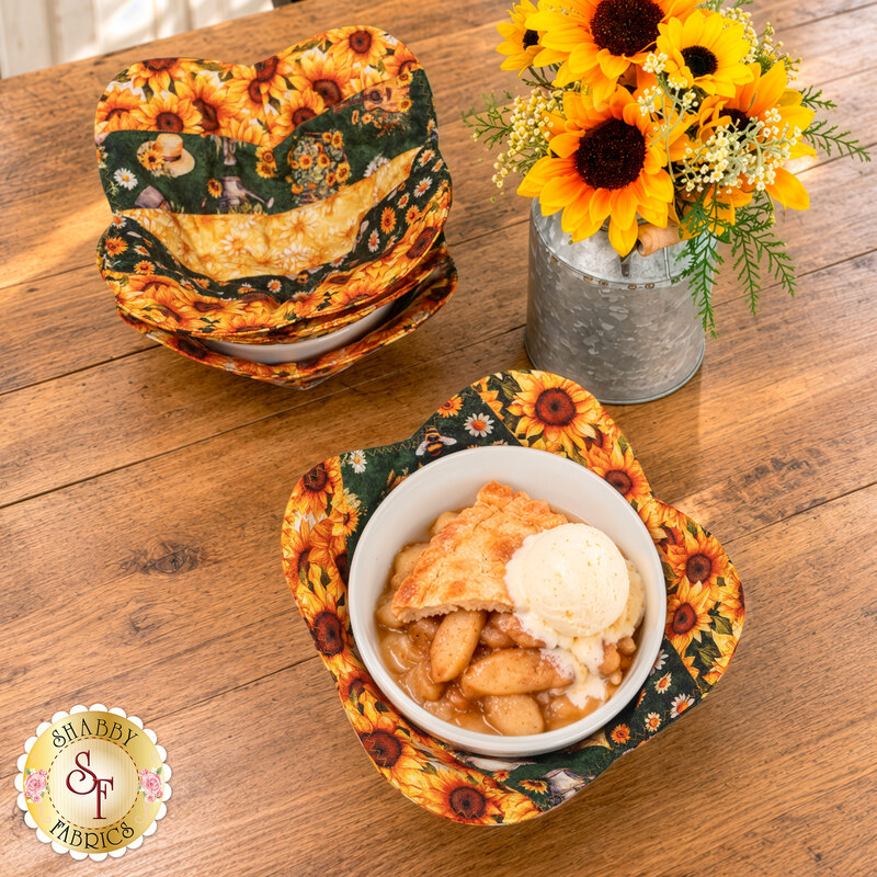 The completed Bowl Cozies, colored in black, yellow, cream, and green fabrics from the Sunflower Serenade collection by QT Fabrics, staged on a wooden counter top with coordinating décor. One of the cozies holds a bowl of apple pie and vanilla ice cream.
