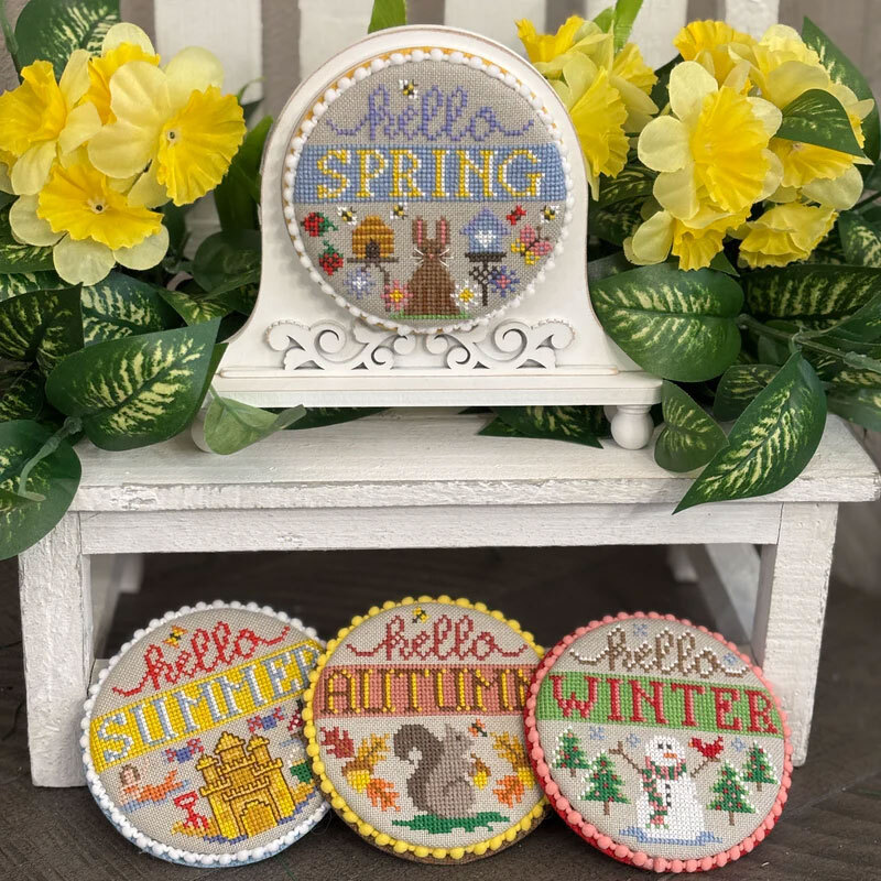 The fully assembled and painted Fleur Mini Round Cross Stitch Frame, staged on a white pedestal framed by yellow flowers, displaying four festive cross-stitch projects.