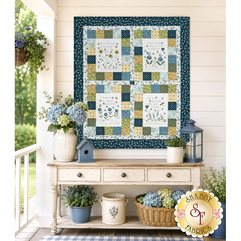 The completed Friendship Garden Wall Hanging, colored in white, navy blue, green, and yellow fabrics from the Friendship Garden collection by Henry Glass Fabrics, hung on a white porch wall and staged with coordinating flowers and décor.