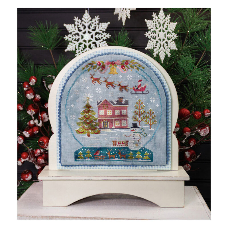 The finished Winter Joy cross stitch project featuring snow globe design with a brick house with a snowman in the yard and Santa flying his sleigh above, framed with decorative trim and displayed on a wooden snow globe frame.