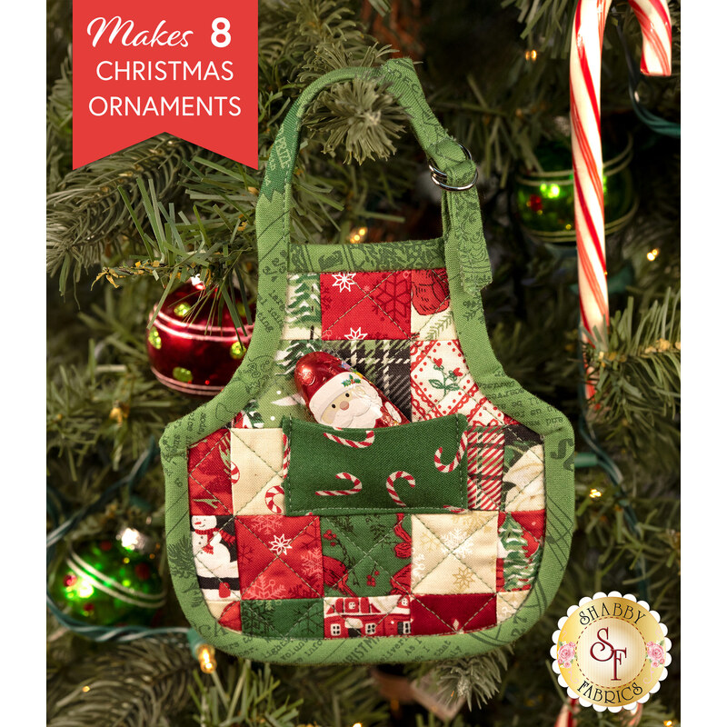 A completed Christmas Apron Ornament, colored in Christmas themed prints from the Holiday Cheer collection by Moda Fabrics, hung on a decorated Christmas tree. A small chocolate Santa peeks out of the dimensional front pocket.