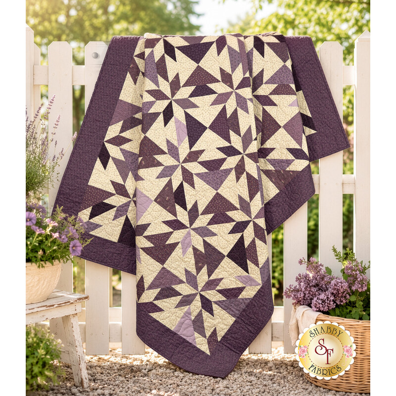 The completed Playing Favorites Quilt colored in rich cream and purple fabrics from the Plumberry Farm collection by Marcus Fabrics, artfully draped over a white fence and staged with coordinating décor.