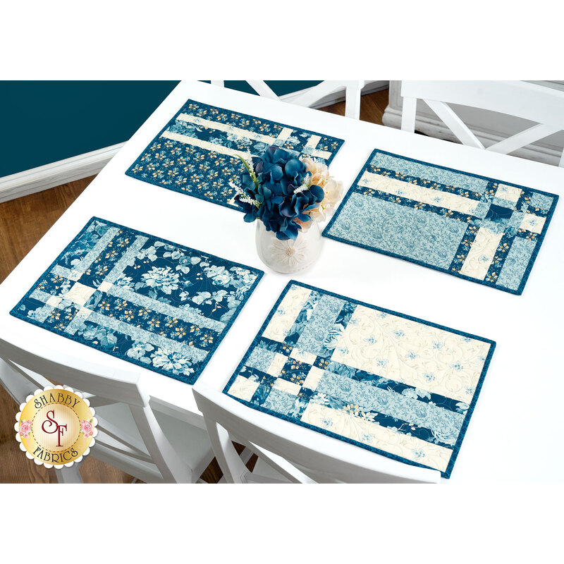 The four completed Fat Quarter Shuffle Mats, colored in cream and blue fabrics from the Special Delivery collection by Andover Fabrics, set on a white table with coordinating flowers.