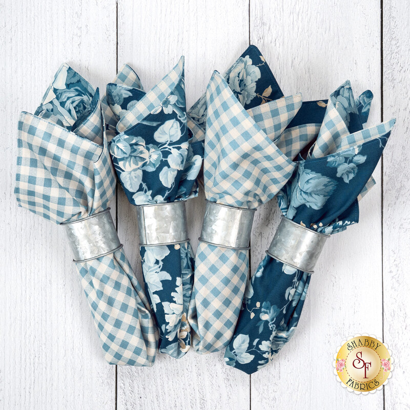 The four completed cloth napkins, colored in cream and blue fabrics from the Special Delivery collection by Andover Fabrics, staged on a white wooden table.