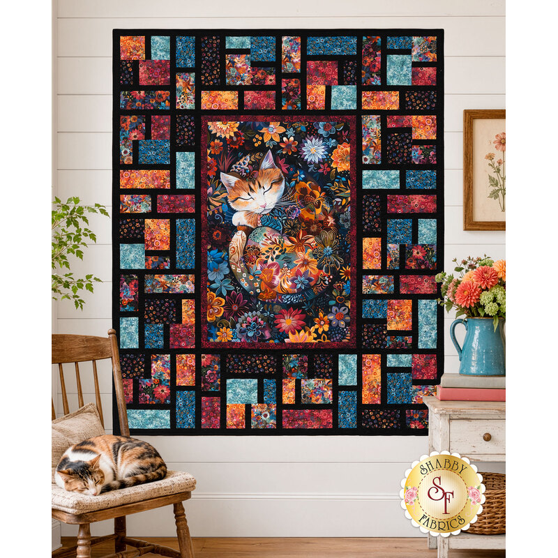 The completed Stained Glass Panel Quilt, colored in orange, magenta, cyan, and black fabrics from the Calico Cat collection by Timeless Treasures, staged on a white shiplap wall with coordinating furniture, décor, and a sleeping kitty cat.