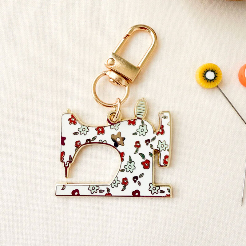 Gold keychain featuring a colorful, decorative enamel sewing machine with floral designs.