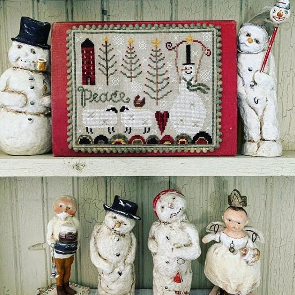Finished Peaceful Snow cross stitch surrounded by vintage winter dolls on a white painted shelf.