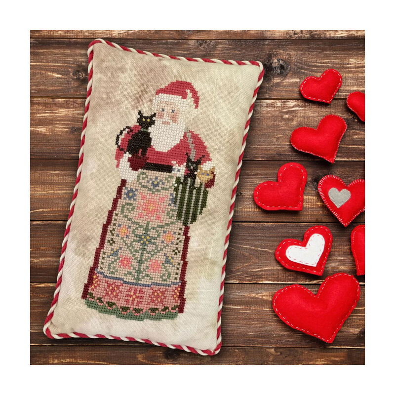 Finished Special Delivery cross stitch pattern of Santa in an apron holding kittens stitched on a pillow placed on a wooden plank backdrop sided by hearts.