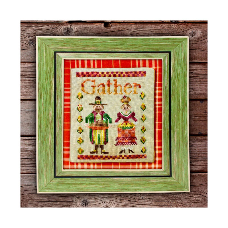 Finished Gather cross stitch pattern with two pilgrims holding Thanksgiving food in a green frame laid on wooden planks.