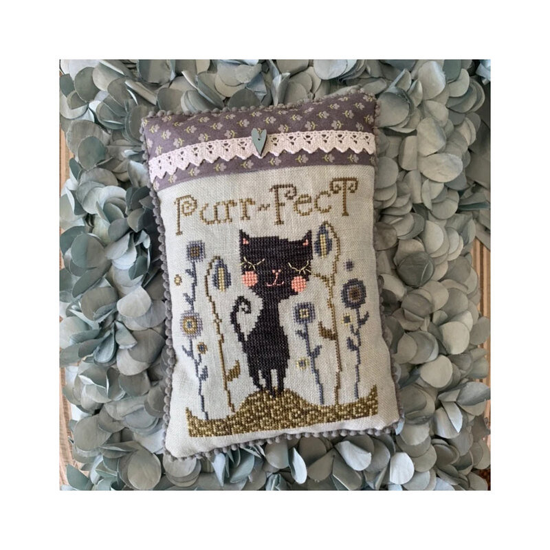 Finished Purr-Fect! cross stitch pattern of a black cat walking through a flower field topped with a pink lace fabric and heart charm stitched onto a pillow.